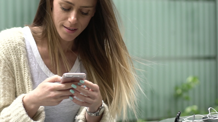 Close-up shot of smiling woman texting on her smart phone while sitting outdoors | Shutterstock HD Video #1034456690
