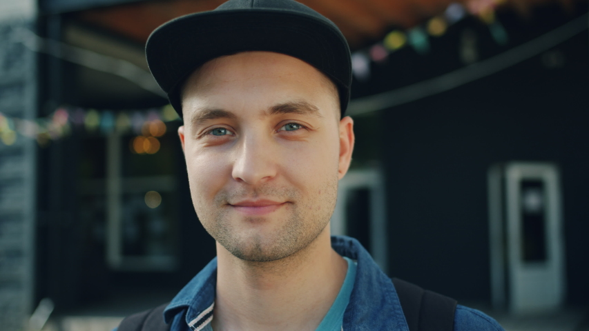 Close-up portrait of smiling young guy wearing trendy cap standing outdoors looking at camera with modern city street in background. People and emotions concept. Royalty-Free Stock Footage #1034469047