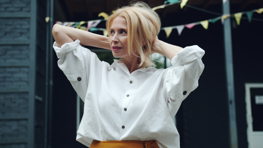 Slow motion of beautiful mature lady touching blond hair then looking at camera smiling standing outdoors in city street. People, beauty and fashion concept.