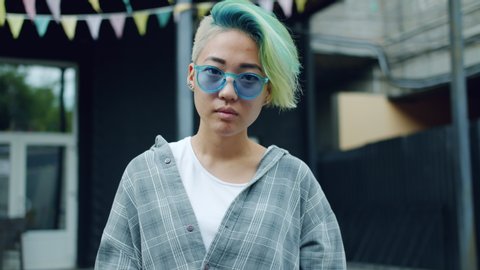 Slow motion of joyful Asian hipster with blue dyed hair and nose piercing raising sunglasses smiling outdoors looking at camera standing in city street alone.