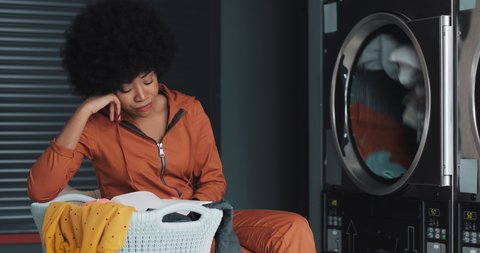 Tired young African American woman reading a book while washing her laundry at laundromat. She yawns and waits for the completion of the process.