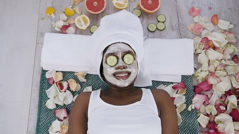 Top view of beautiful young african woman in white towel on head lying with pieces of cucumber on her face in beauty salon. Beautiful young woman getting moisturizing facial mask at spa centre. Beauty