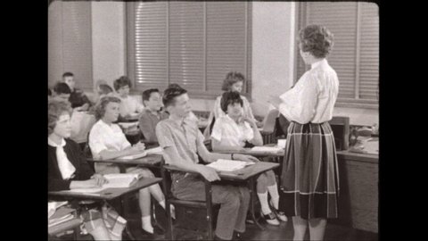 1940s: Woman stands in front of classroom, speaks, students raise hands, lower them, boy speaks, more students raise hands. Girl with high bangs speaks. Woman holds book in front of chalkboard.