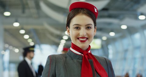 Close up of the attractive young Caucasian brunette stewardess in uniform looking and smiling to the camera in the airport. Portrait.