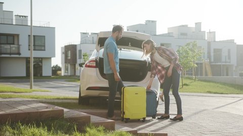 Young affectionate parents packing things into a car trunk to go on vacation. Happy teenage girl dancing around in a inflatable lifebuoy expecting a fun roadtrip. Summer.