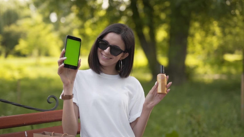 Caucasian Girl With Sunglasses Sitting on Park Bench and Showing With Smile to Camera Green Layered Smartphone and Spay Bottle. Light Camera Movement