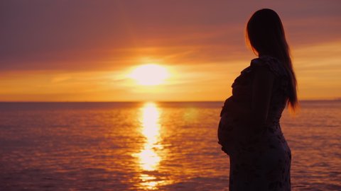 Young pregnant woman with a big belly looks at the majestic sunset over the sea. In anticipation of a child