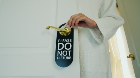 Unrecognizable woman in a white house coat hangs a sign on the door Do not disturb
