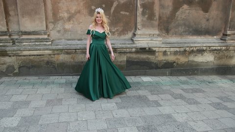 spectacular girl, blonde, in green dress with deep neckline, with a chic hairstyle and bright make-up, crown on her head, posing and smiling in front of camera, near the old building, slow shooting