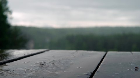 Raindrops on wooden table on background of green forest. Stock footage. Cloudy rainy weather hung over park and rain drops hit picnic table