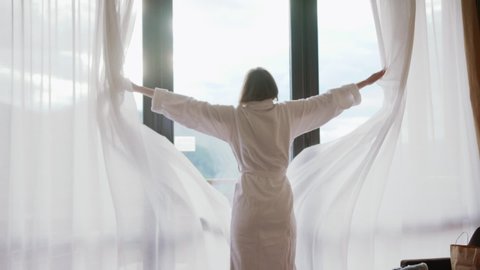 Close up back view young woman opening curtain lace standing in luxury apartment home or hotel looking through window enjoying wellbeing light city skyscrapers morning close up slow motion