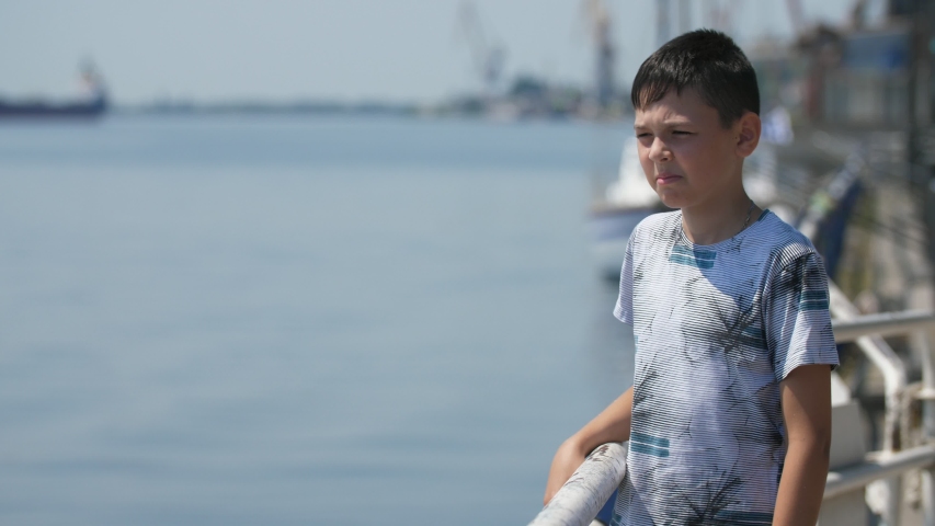  Cheerful view of a happy brunette boy with a short haircut in colorful T-shirt standing, smiling on Dnipro embankment on a sunny day in summer in slo-mo
