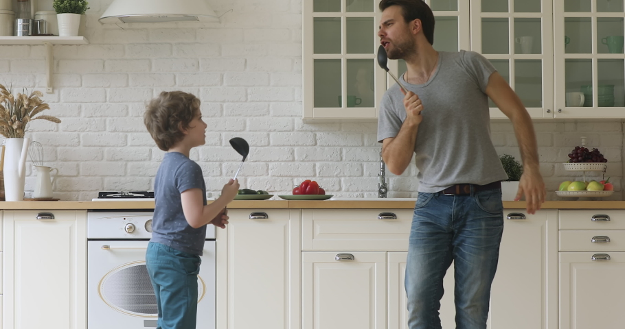 Funny artistic father and cute little kid son singing song in kitchen, happy family dad with child boy holding spoon ladle kitchenware microphones enjoy karaoke dancing having fun in kitchen together Royalty-Free Stock Footage #1034586188