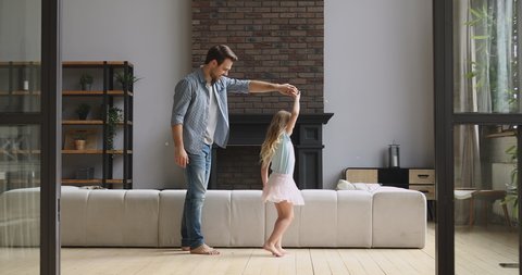 Loving young dad stand on knee holding kissing hand dancing with cute little child daughter princess in modern living room, happy father and small kid girl playing enjoy sweet time together at home