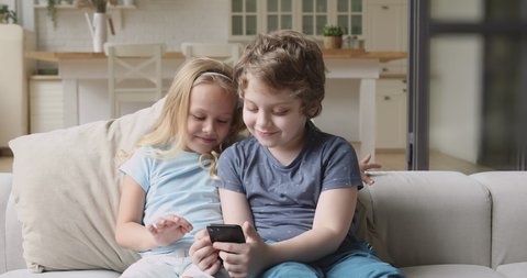 Two funny little kids siblings holding phone using smartphone playing mobile game browsing social media taking selfie at home having fun with cellphone device sit on sofa, children and gadget concept