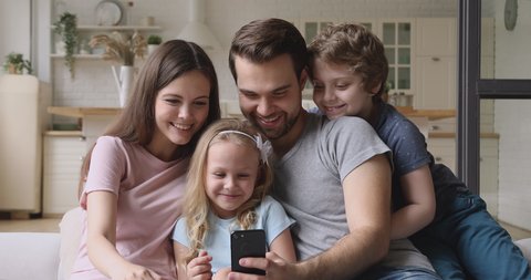 Happy family parents with cute little kids children son daughter sit on sofa laugh having fun using smart phone funny social media app, take selfie, watching cartoons look at cellphone screen at home