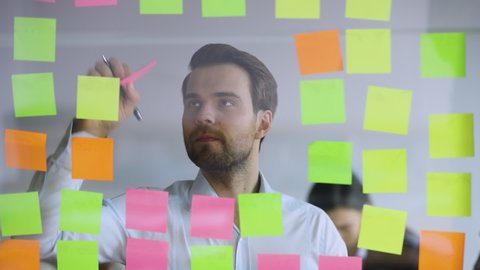 Male corporate leader scrum master write new project tasks on post it sticky notes put reminders planning work organization developing strategy attaching replacing stickers on glass board in office