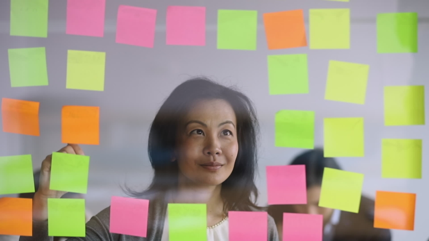 Serious mature female asian manager checking information on scrum board, focused mid aged corporate leader writing tasks ideas list on post it sticky notes attaching stickers to glass wall in office Royalty-Free Stock Footage #1034587580