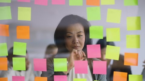 Serious mature female asian manager checking information on scrum board, focused mid aged corporate leader writing tasks ideas list on post it sticky notes attaching stickers to glass wall in office