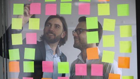 Two happy businessmen colleagues team write on post it sticky notes look at glass wall scrum board brainstorm on work organization task management discuss strategy plan in office at teamwork meeting