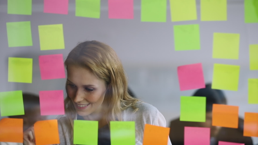 Young businesswoman office worker replacing sticky notes on planning board, female corporate leader write project tasks on post it sticky notes organize work attaching stickers on scrum glass wall Royalty-Free Stock Footage #1034587589
