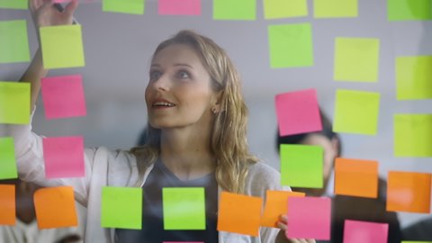 Young businesswoman office worker replacing sticky notes on planning board, female corporate leader write project tasks on post it sticky notes organize work attaching stickers on scrum glass wall