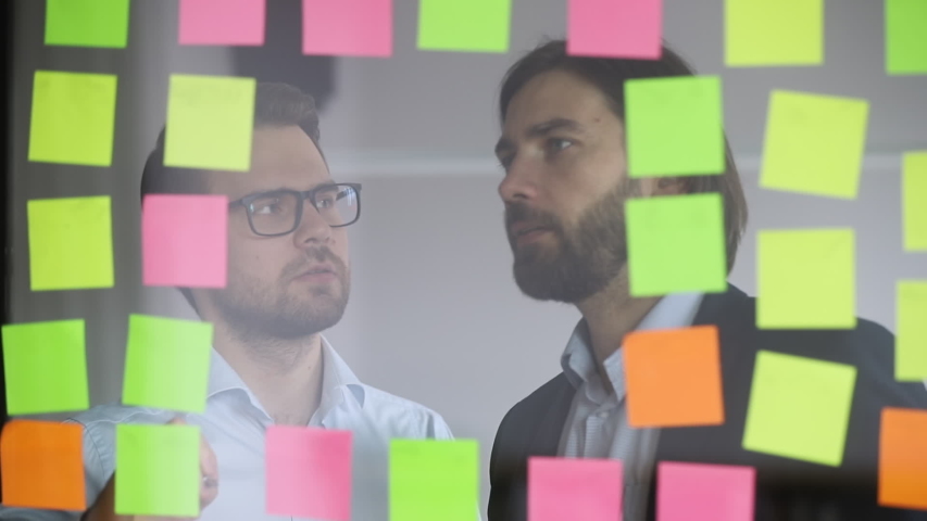 Two focused male colleagues partners team writing business ideas on paper post it sticky notes on glass wall discussing teamwork brainstorm on strategy work plan in corporate office at scrum meeting Royalty-Free Stock Footage #1034587664
