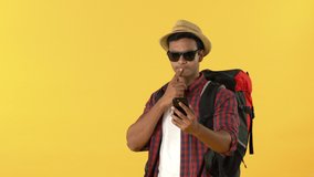 Young happy Indian tourist man with backpack smiling and waving bye while having video call on mobile phone against yellow background