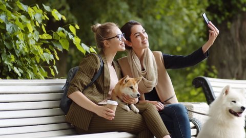 Lockdown of mixed-race young woman holding smartphone and making selfie with her female friend and Shiba Inu lying on her knees while Samoyed sitting nearby. Then women looking through photos