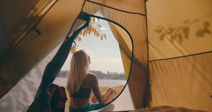 Happy traveler woman with open arms sitting near tent camp around mountains lake under sun light enjoying the leisure and freedom. 4K slow motion video