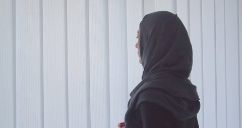 Closeup portrait of young attractive muslim woman in hijab looking through the window with tulle indoors in apartment