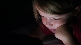 Close up of face and hands of little girl playing computer games lying in his bed in night. Child using tablet pc.
