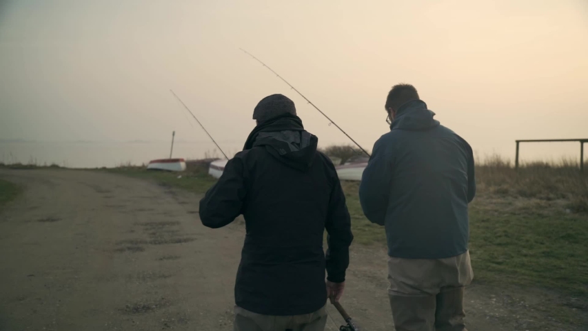 two men walking with fishing poles in sunrise and water and boats in distance