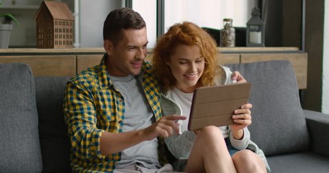 Happy young couple sitting on sofa, using tablet computer, browsing, laughing at funny photos, videos, having fun together, enjoying their weekend at home, Caucasian. 4K, shot on RED camera.