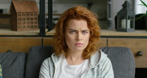 Portrait of young woman changing mood from sad to happy and positive, sitting on sofa at home, smiling, looking at camera, full joy, red curly hair, blue eyes, Caucasian. 4K, shot on RED camera.