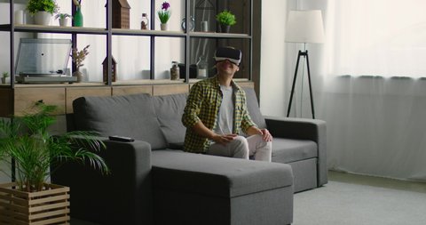 Young man is wearing virtual reality headset, using gestures to control images, rips and sniffs virtual fruits, enjoys 360 video imagination concept, sitting on sofa. 4K, shot on RED camera.