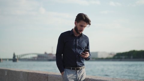 Zoom out slow motion shot of young businessman standing on embankment near river with hand in pocket, looking away thoughtfully and text messaging on cell phone