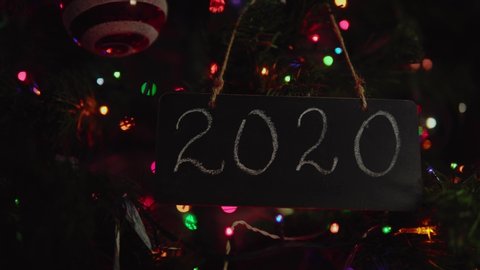 Black letter board with number 2020 on it hanging on a branch is Christmas tree among glass balls and multicolor round bokeh in background. Camera approaches chalkboard, backlight flashes.