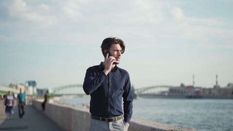 Slow motion panning shot of young businessman talking on cell phone while walking on embankment near river. Man ending phone call, stopping and looking at view thoughtfully