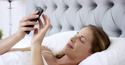 beautiful woman using phone in bed managing smart connected home with mobile app