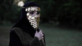 pretty woman with bright makeup and expressive eyes wears golden tribal face veil and stands against forest slow motion
