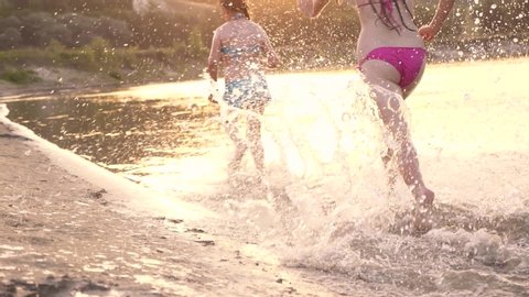 happy kids running along the beach. Girls run on sand along shore on beach splashing water drops and laughing. Happy and free teenagers on a summer vacation have rest at sunset. Teamwork