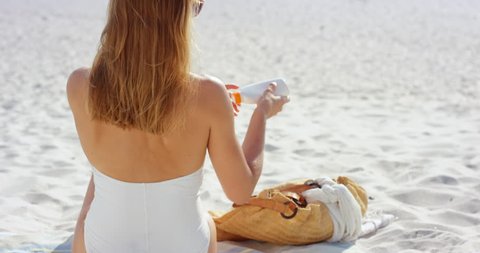 beautiful young woman applying sunscreen to arms on beach wearing white swimsuit