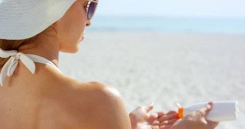 Close up of woman applying sunscreen to shoulder on tropical beach wearing one piece white bathing suite and white hat