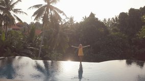 Drone aerial view of Young woman arms outstretched on the edge of an infinity pool in a rainforest. Girl enjoys freedom on holidays breathing fresh air in pure nature 
