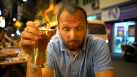 Drunk man drinking beer and talking to camera, sitting in bar