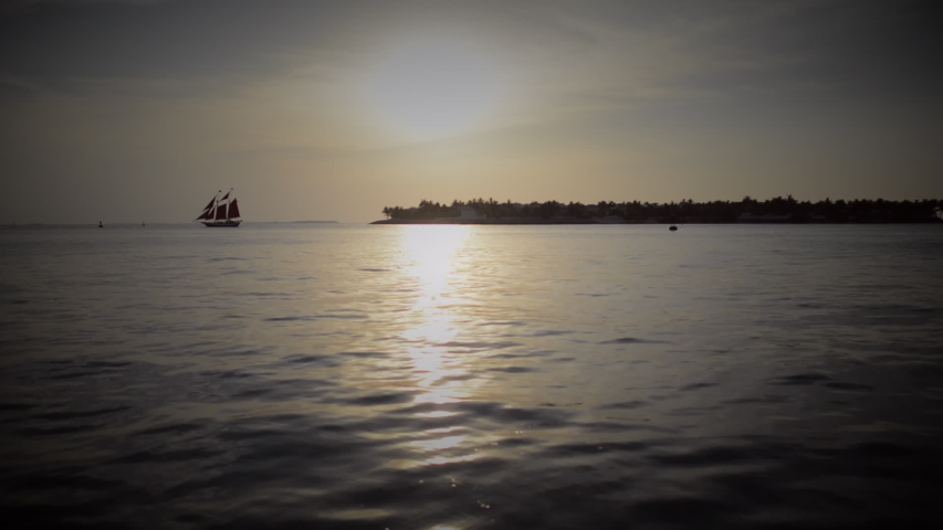 Beautiful evening view from shore on calm sea, sunset and small sailing boat with red sails near island. Sun rays fall and reflect on the water. Key West, Florida, USA