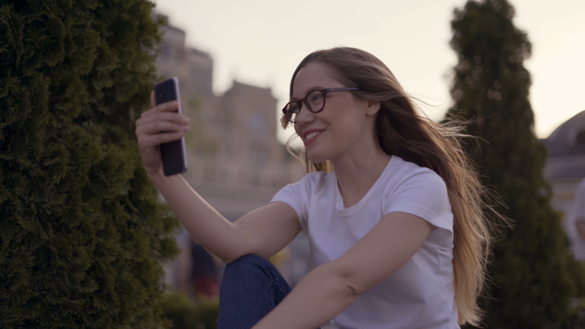 Hello there! Young girl is waving to the screen to her boyfriend in camera while having video call in the spring park outdoors, sitting on the bench 4K