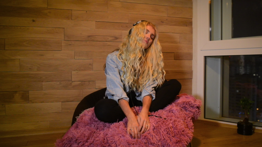 Young blonde woman siting in corner near window in headphones and listening music. Beautiful girl gesticulating and singing in evening time at home.