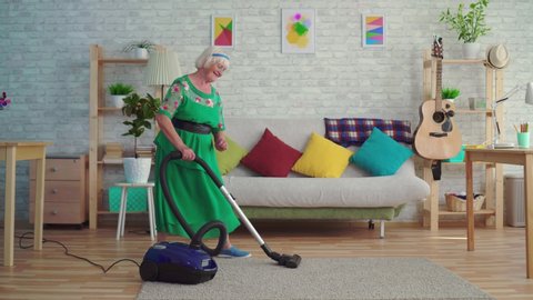 Cheerful happy old woman with gray hair vacuuming the carpet in the living room and dancing